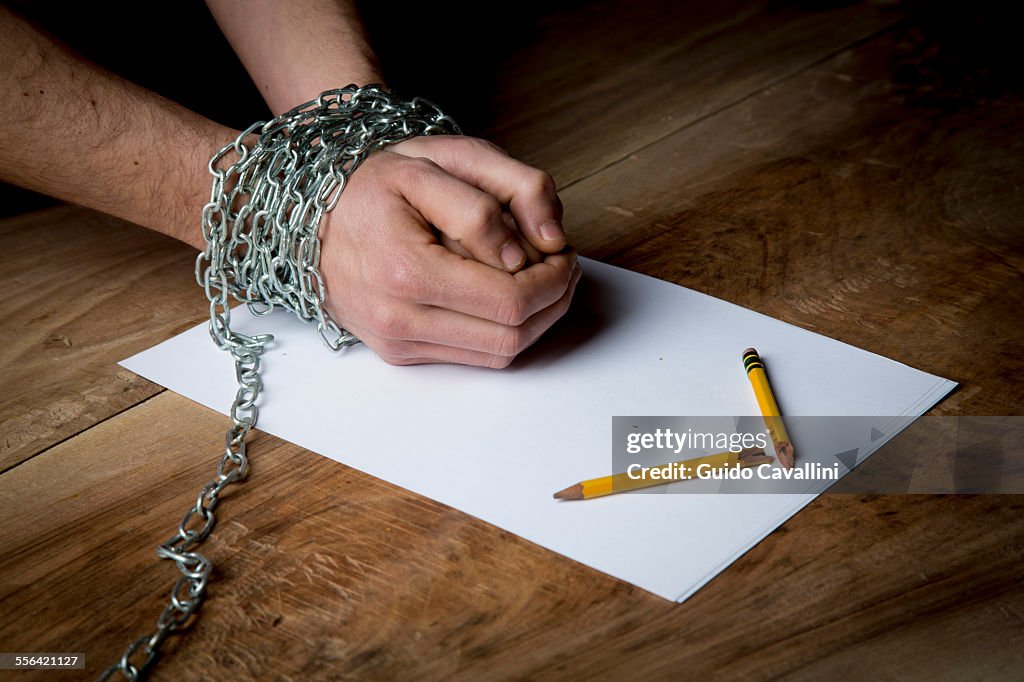 Young man with chains wrapped around his wrists, paper and broken pencil in front of him, focus on hands