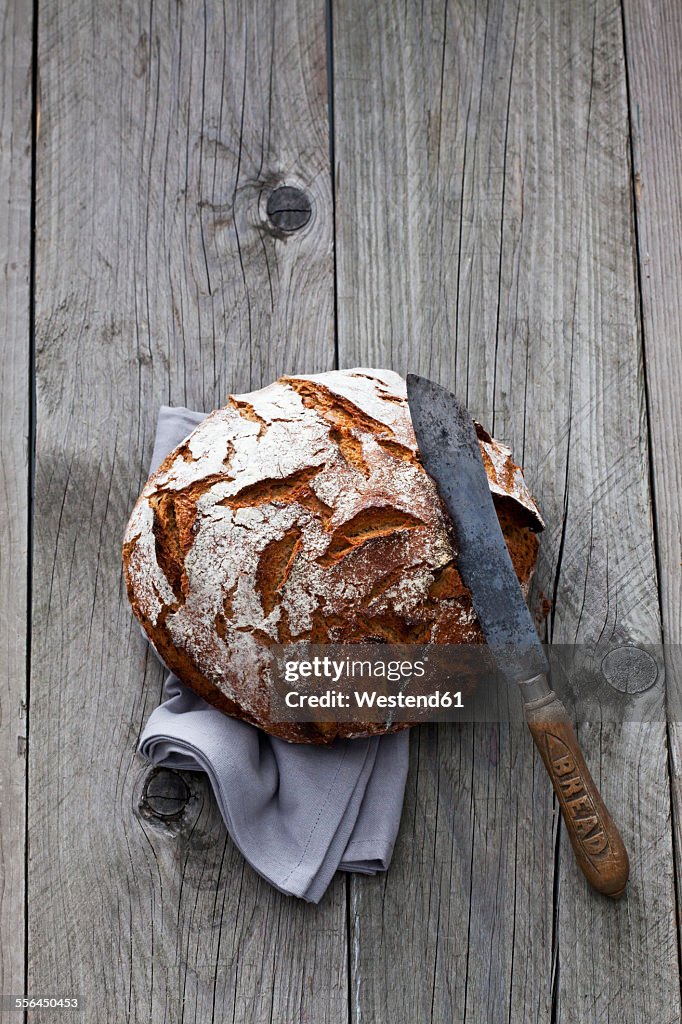 Round crusty bread, old bread knife and kitchen towel on wood