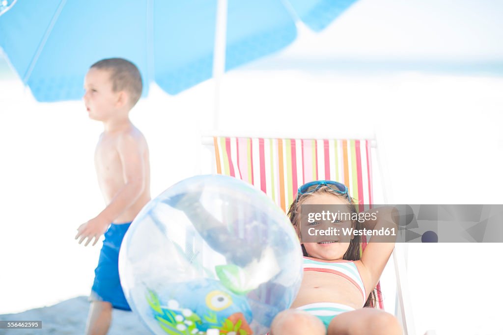 Smiling girl on beach relaxing on a beach chair with boy passing by