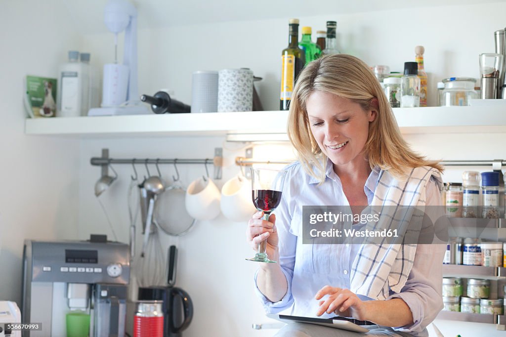 Smiling woman in kitchen drinking glass of red wine and using digital tablet