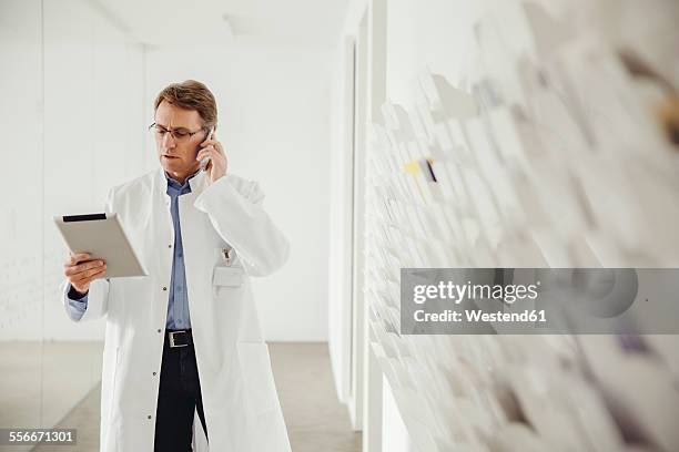 mature man in lab coat on phone looking at digital tablet - doctor lab coat stock-fotos und bilder