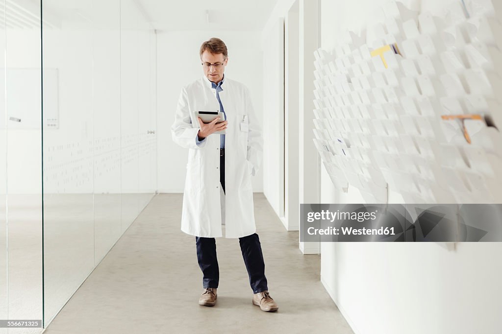 Mature man in lab coat standing in hallway looking at digital tablet