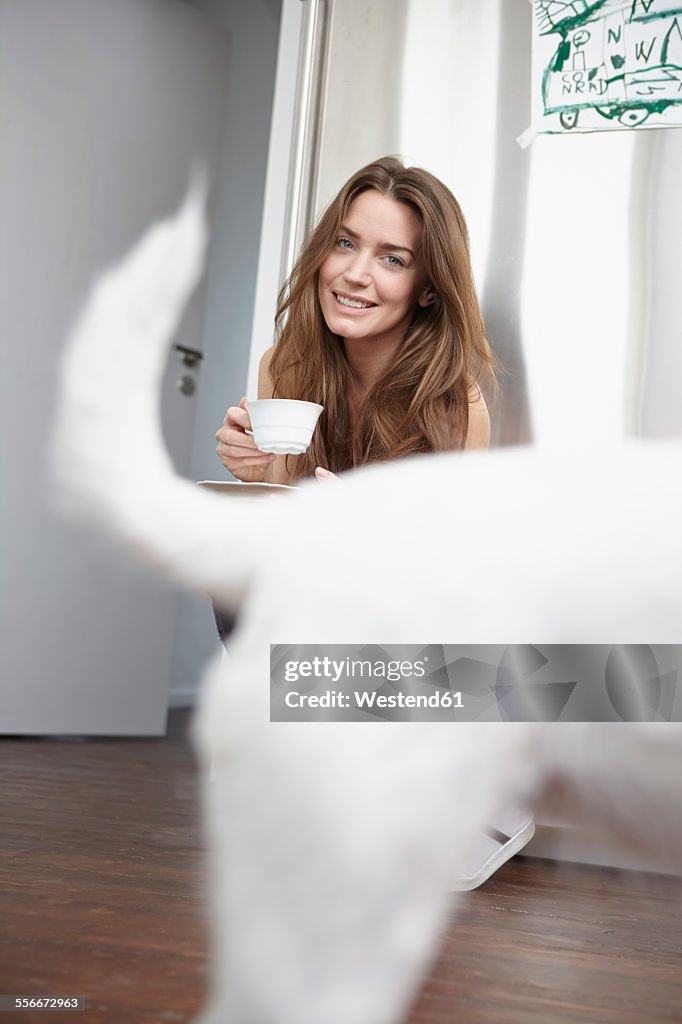 Young woman in kitchen drinking cup of coffee, dog in foreground