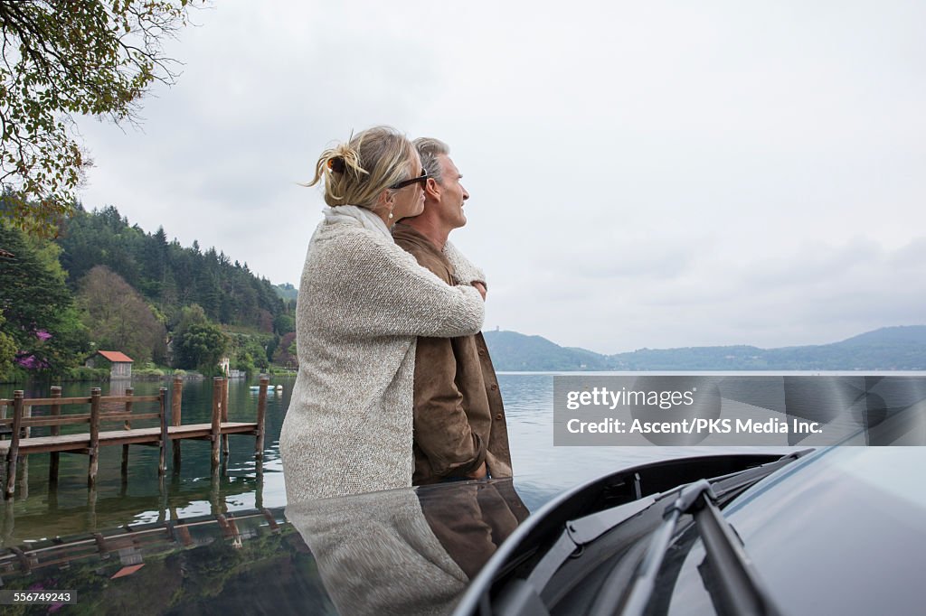 Couple embrace at edge of lake, beside car