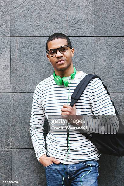 portrait of young man with green headphones and backpack leaning at house front - auf den schultern stock-fotos und bilder
