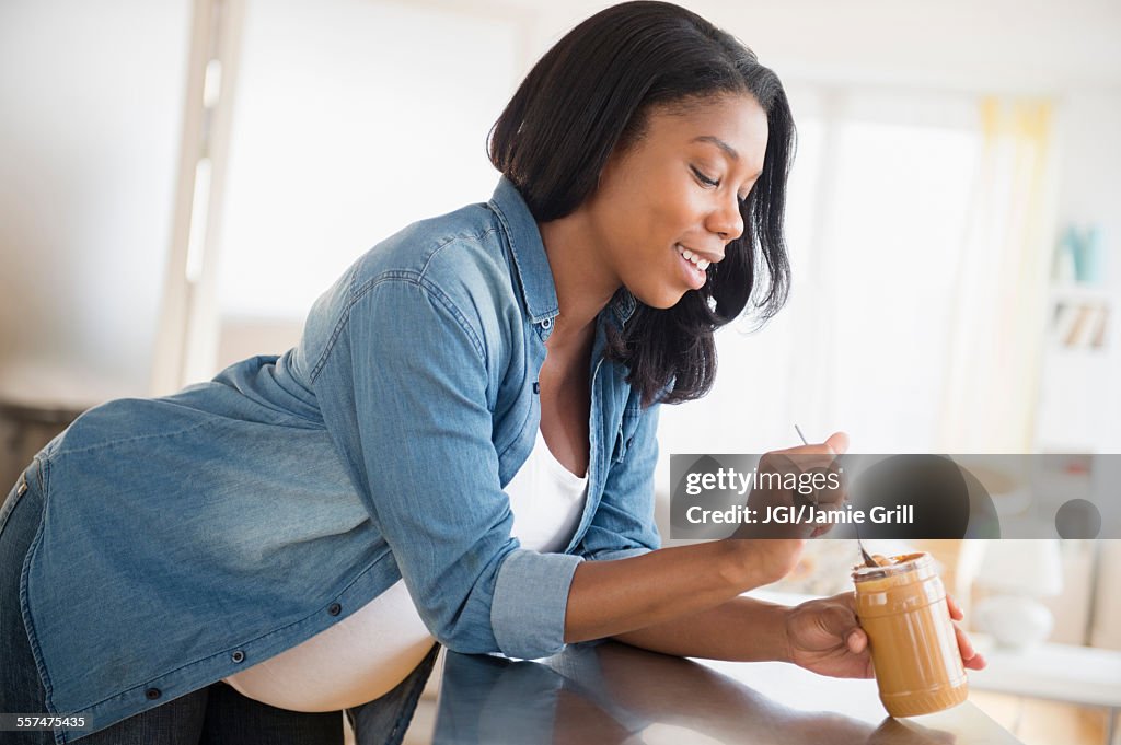 Black pregnant woman eating peanut butter in kitchen