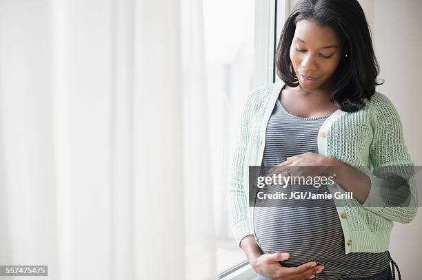 black pregnant woman holding her stomach at window - abdomen humano fotografías e imágenes de stock