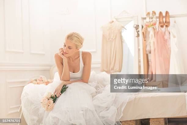 pensive bride sitting with bouquet on bed - wedding dress photos et images de collection