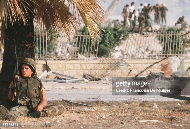 An American sits under a palm tree with his M16 as, in the background, comrades inspect the damage after the suicide bombing that destroyed the...