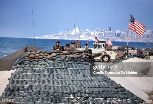 An American Marine holds binoculars as he looks out from a defensive position reinforced by many sandbags in front of several vehicles which fly the...