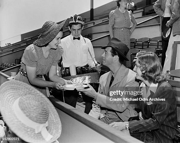 Actor Robert Taylor and his wife actress Barbara Stanwyck attend a game as they are offered food and a Coca-Cola in Los Angeles, California.