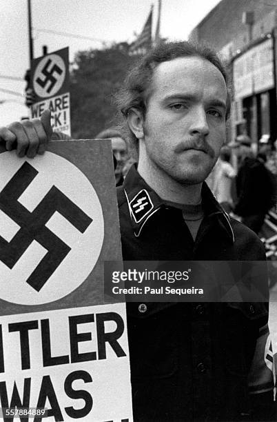 Portrait of an unidentified neo-Nazi, dressed in a shirt adorned with SS collar insignia, as he holds a sign across the street from Harry S Truman...