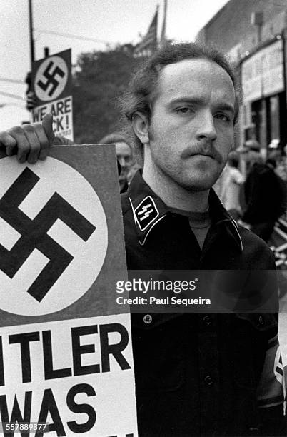 Portrait of an unidentified neo-Nazi, dressed in a shirt adorned with SS collar insignia, as he holds a sign across the street from Harry S Truman...