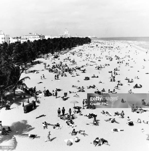 An expansive vista of vacationers and tourists as they enjoy the sun and surf at Miami Beach, Florida, mid 20th Century. In the distance hotels are...