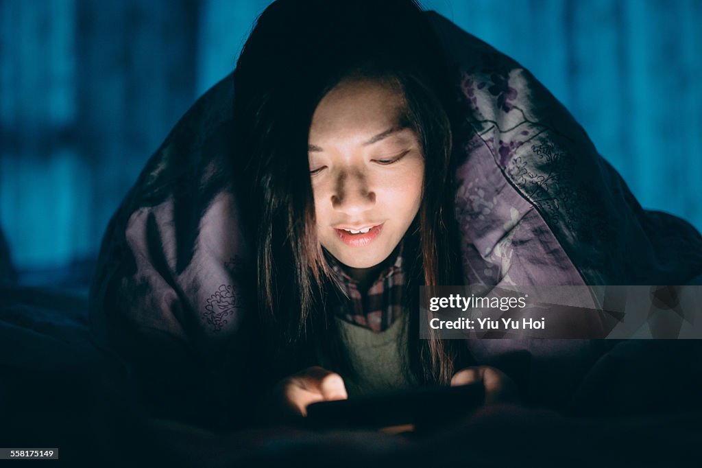 Woman texting on smartphone under the bed sheets