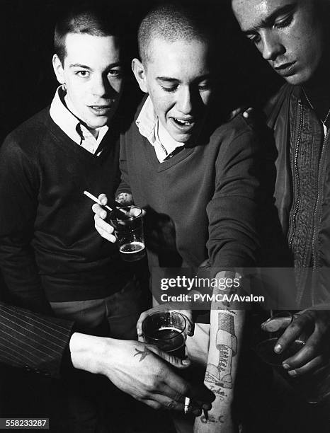 Three skinheads, drinking, showing a 'Kill Mods' tatoo, UK, 1980's.