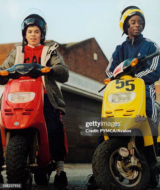 Two teenagers sitting on their mopeds one red one yellow outside a garage.