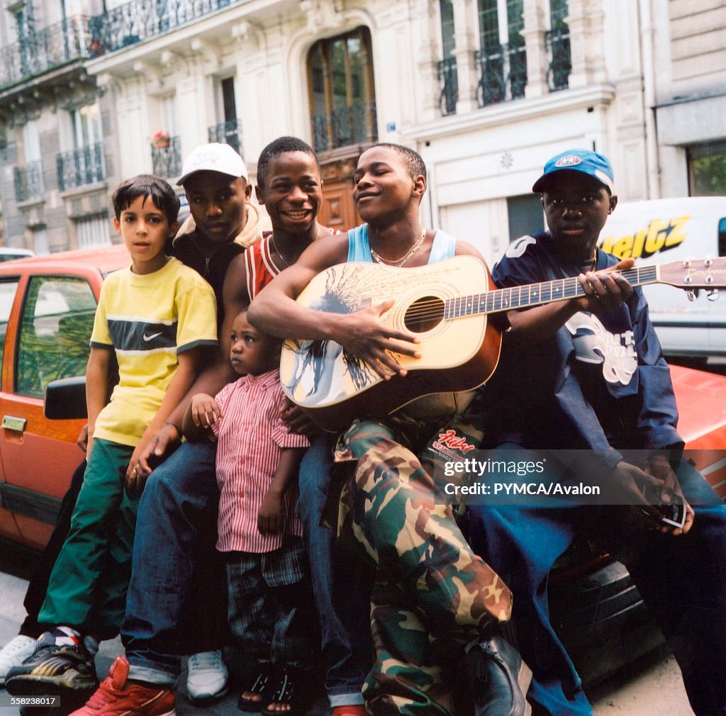 Group of teenagers sitting on a car in Paris one boy playing a guitar