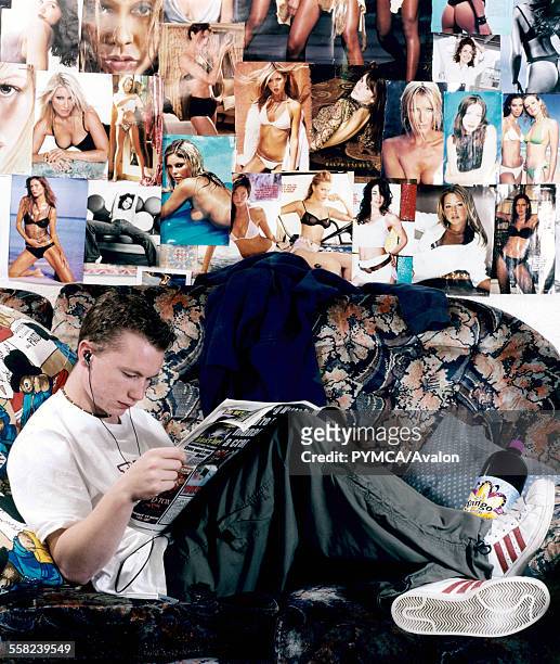 Teenage boy listening to music and sitting on a sofa reading the newspaper surrounded by posters of naked women.