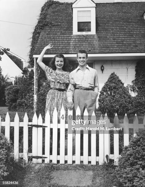 Young couple smiling and waving from behind the white picket fence of their suburban home, 1947.