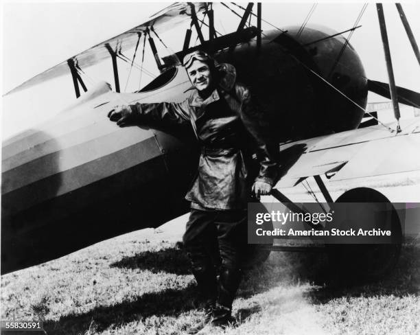 Portrait of a pilot wearing a leather jacket and goggles smiling and leaning against a World War I war plane, 1910s