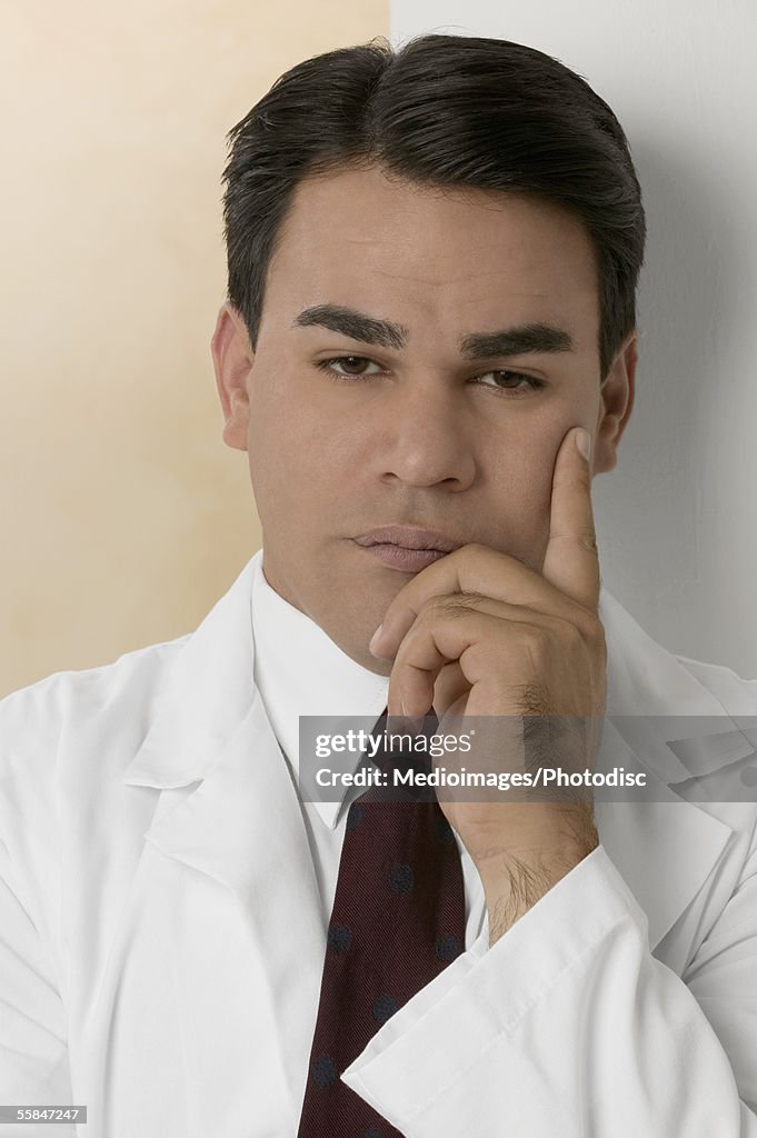 Pensive male doctor leaning against wall with hand on chin