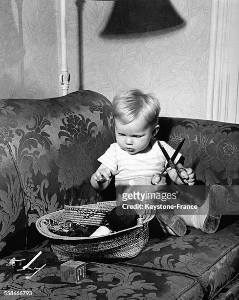 Un tout jeune garçon assis sur un canapé joue avec une paire de ciseaux trouvée dans un panier laissé et en évidence et contenant un nécessaire à...