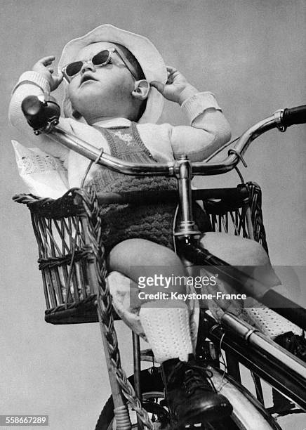 Un jeune enfant avec des lunettes noires tient son chapeau de soleil à deux mains sur le porte-bagages d'un vélo, le 4 octobre 1954.