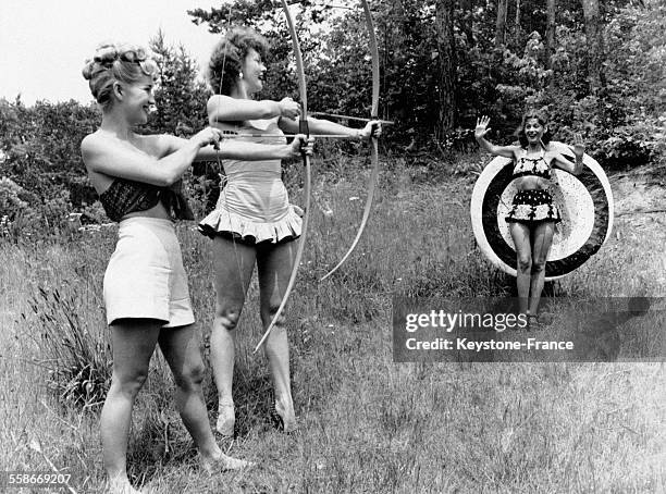 Deux jeunes femmes munies chacune d'un arc s'apprêtent à tirer une flèche sur une cible devant laquelle se tient une troisième jeune femme.