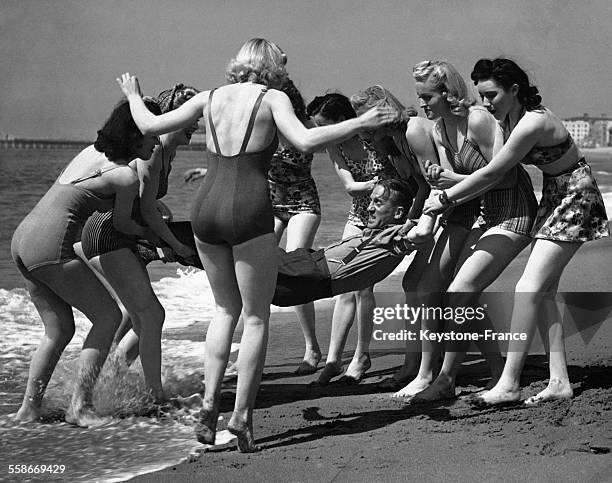 De jeunes femmes en maillot de bain porte un homme tout habillé pour le mettre à l'eau.