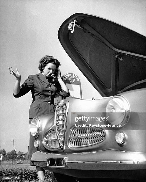 Une femme près de sa voiture au capot ouvert passe un appel téléphonique grâce à une borne téléphonique d'urgence sur le bord de la route.