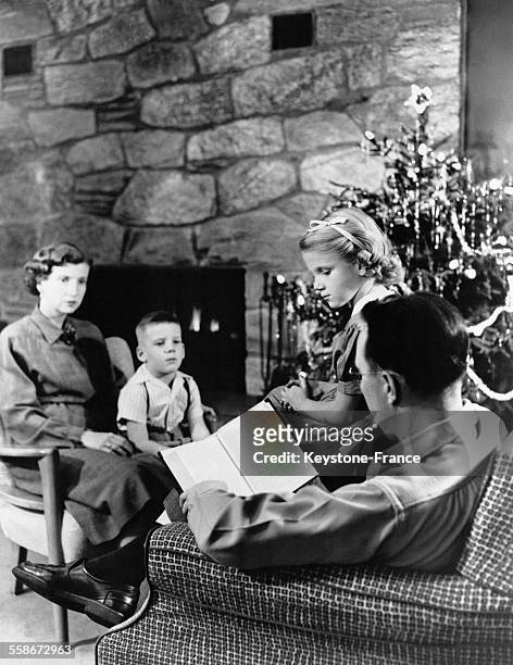 Un père de famille assis dans un fauteuil lit un livre à ses deux enfants et à sa femme rassemblés autour du sapin de Noël.