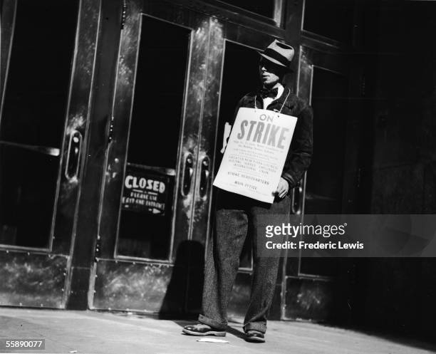 Striker stands outside the closed doors of a building and wears a sign around his neck which reads 'on strike,' New York, New York, 1930s. A sign in...