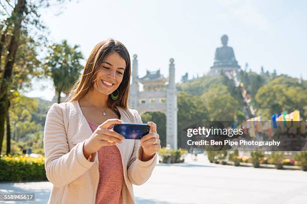 young female tourist reviewing smartphone photographs in front of tian tan buddha, po lin monastery, lantau island, hong kong, china - großer buddha stock-fotos und bilder