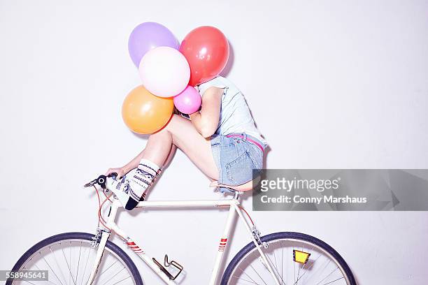 studio shot of young woman sitting on bicycle hiding behind bunch of balloons - hidden object stock pictures, royalty-free photos & images