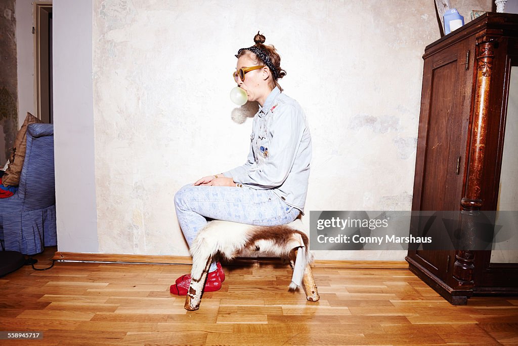 Young woman sitting on hassock wearing sunglasses and blowing bubblegum