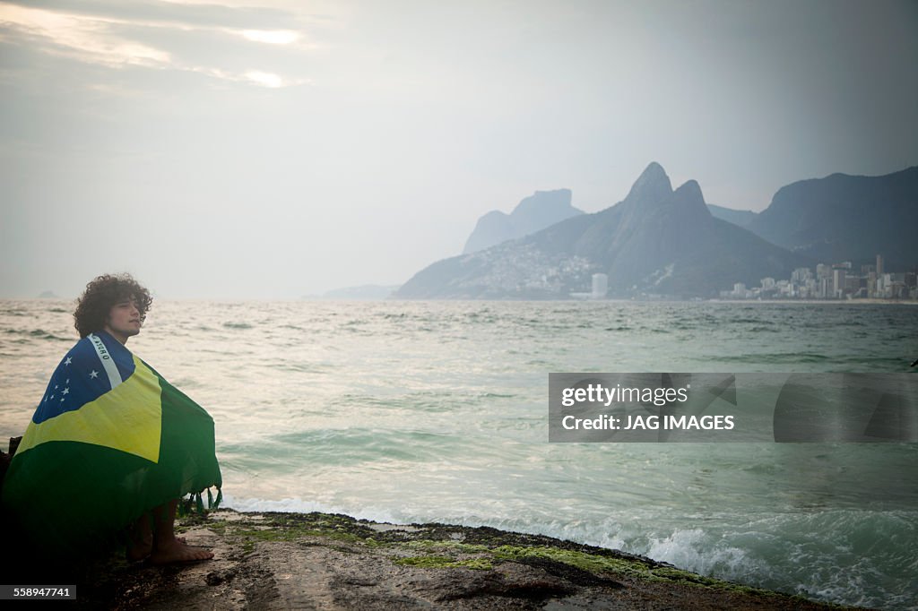 Young man wrapped in Brazilian flag sitting on rock, Ipanema beach, Rio De Janeiro, Brazil