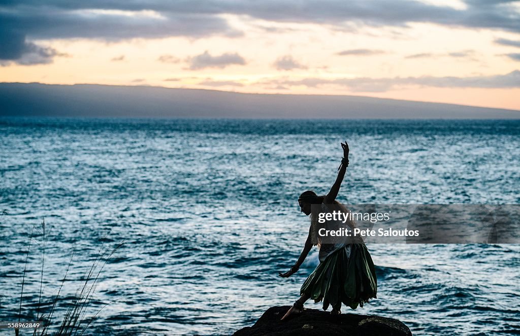 Silhouetted woman hula dancing on coastal rocks wearing traditional costume at dusk, Maui, Hawaii, USA