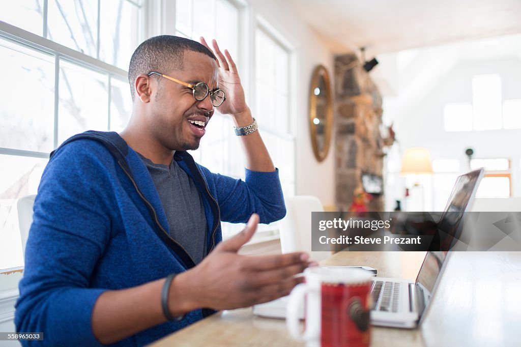 Mid adult man with hands raised in disbelief using laptop at kitchen counter