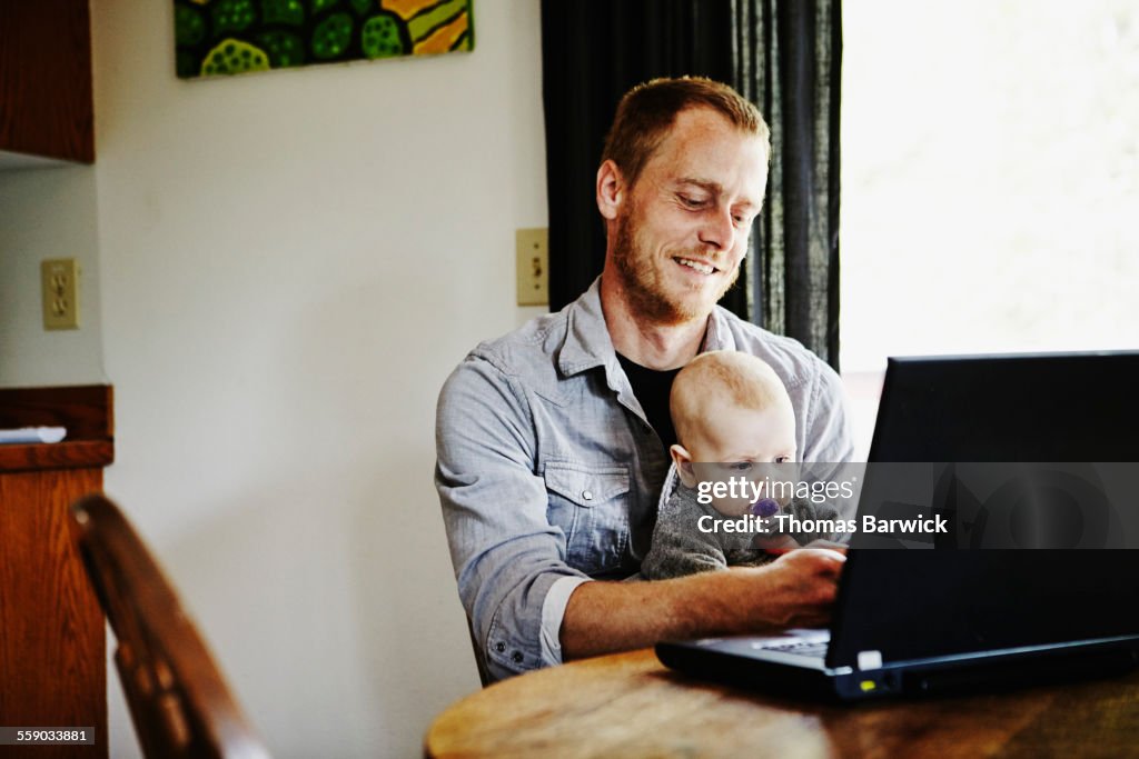 Father sitting working on laptop with baby on lap