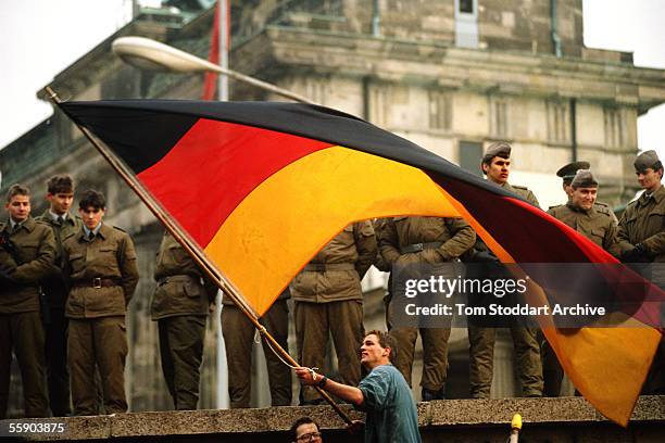 Jublilant man waves the German national flag under the gaze of suspicious East-German border guards on top of the Berlin Wall on the morning of...