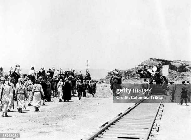 Camera crew, with camera crane mounted on railway track, films a scene of actors walking and riding camels through the sand on the set of the film...