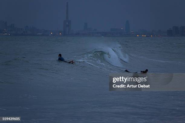 Guys practicing surf at dusk in the Barcelona's shoreline during the spring storms with the city lights on background. Masnou, Catalonia, Europe.