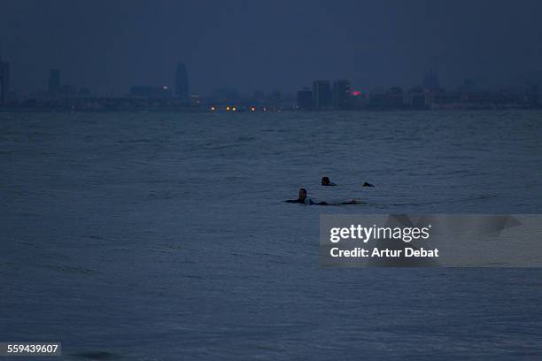 Guys practicing surf at dusk in the Barcelona's shoreline during the spring storms with the city lights on background. Masnou, Catalonia, Europe.