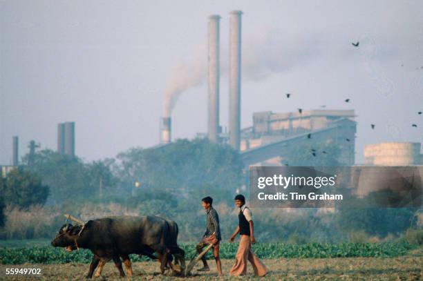 Farmers ploughing field using traditional method next to polluting industrial scene, India.