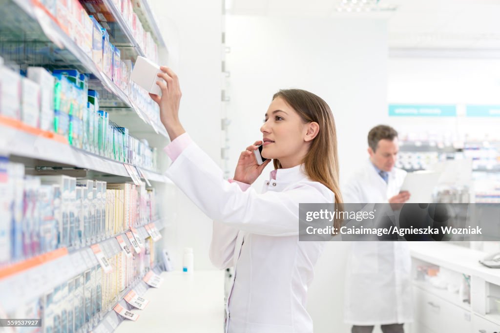 Pharmacist talking on cell phone and removing box from shelf in pharmacy