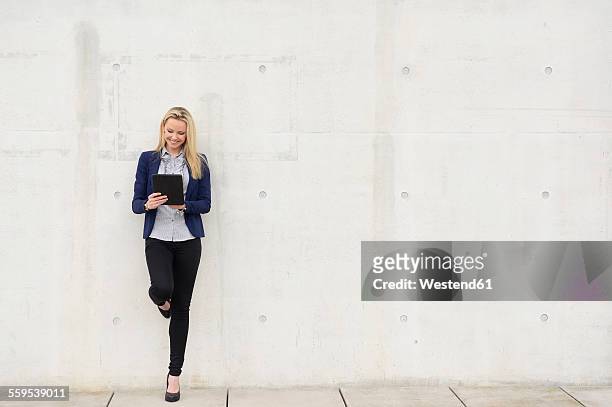 smiling businesswoman with digital tablet leaning on concrete wall - blonde long legs 個照片及圖片檔