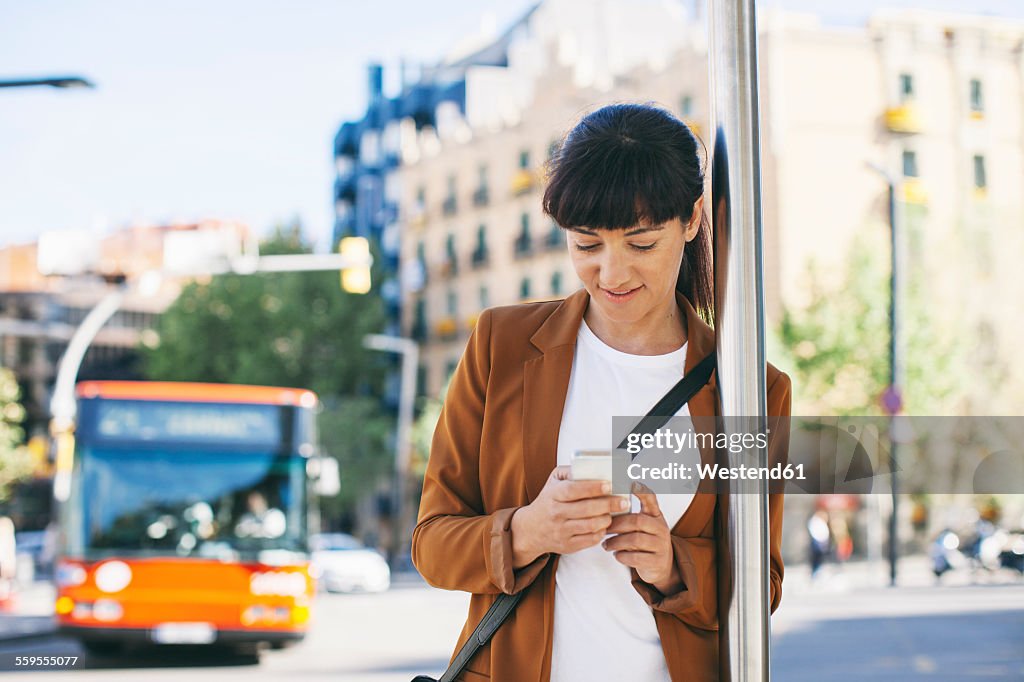 Spain, Barcelona, businesswoman with smartphone waiting at the bus stop