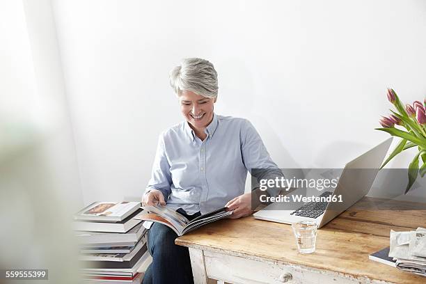 mature woman sitting at table with magazine and laptop - glass magazine stock pictures, royalty-free photos & images