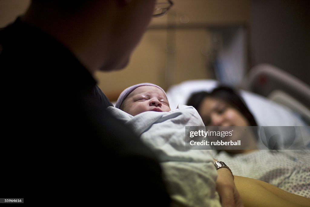 Father carrying his newborn baby girl with his wife lying on a bed